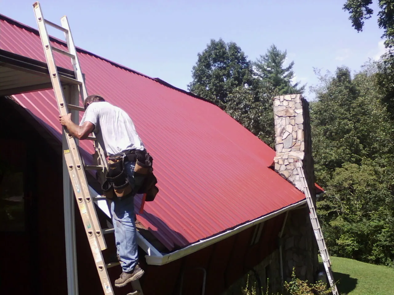Roofer on a ladder beside a red metal roof and stone chimney — Soffit & Fascia Repair by Roofing Brothers in Dawson